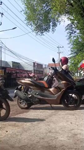 The video shows a street scene with several motorbikes parked on the side of a dirt road. In the foreground, a bronze-colored scooter is prominently featured, with a person wearing a red helmet sitting on it. The person appears to be looking to the right. Behind the scooter, other motorbikes and a few people are visible. In the background, there are shops with awnings and trees lining the street, suggesting an outdoor commercial area. Power lines are strung across the upper part of the frame, and the sky is visible. The lighting indicates it is daytime, likely sunny.