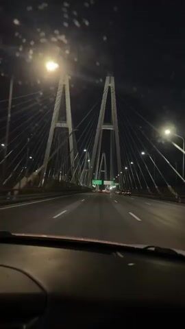 The video depicts a nighttime scene from the inside of a car driving on a bridge. The bridge is a cable-stayed bridge with two prominent towers. The road is illuminated by streetlights and the headlights of other vehicles. The sky is dark, and the windshield of the car has some water droplets on it. The car is moving forward, and other cars are visible on the road.