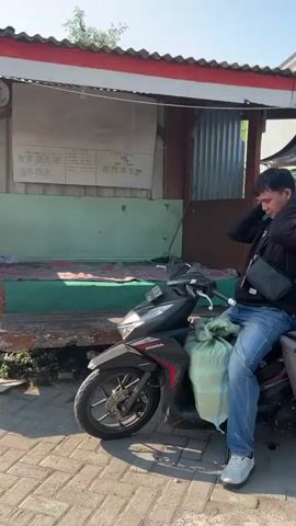 A man in a black jacket and blue jeans is sitting on a black motorcycle. He is adjusting something around his neck. There is a large green bag tied to the back of the motorcycle. The motorcycle is parked on a paved surface, possibly a sidewalk. In the background, there is a small building with a red and white striped awning and a sign on the wall. The lighting suggests it is daytime.