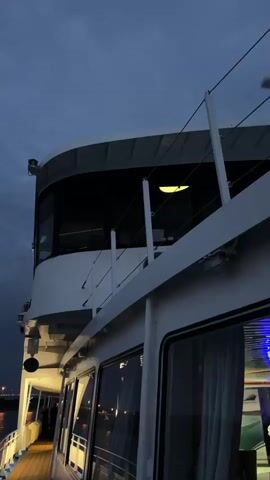 The video shows a close-up, low-angle shot of a white ferry or boat at dusk. The sky is dark blue with some clouds. The boat has a multi-level structure with windows. The upper level appears to be the bridge, with a large, curved window showing a yellow light inside. The lower level has a deck with wooden planks and several windows with curtains. Metal railings and cables run along the side of the boat. The scene suggests the boat is docked or moving slowly in a dimly lit area, possibly a harbor, with faint lights visible in the distance.