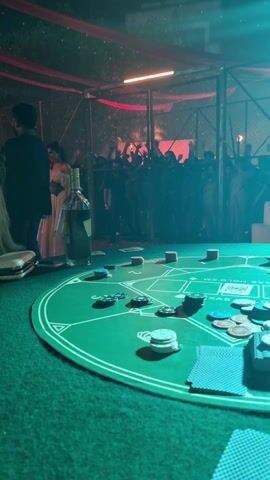 The video shows a film set at night, lit with bright lights and red decorations hanging from the ceiling. Several people are present, some operating cameras and other filming equipment. In the initial shot, a poker table is in the foreground with cards and chips scattered on it, along with a bottle of liquor. The background shows more people silhouetted against the lights, some with their hands raised. The camera moves around to capture various angles of the set and the film crew in action.