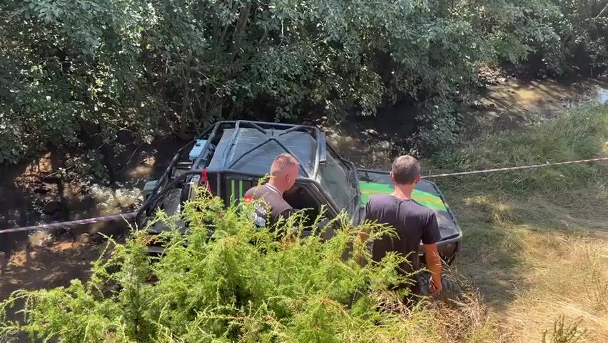 In a wooded area with a small stream, two men are observed next to a modified off-road vehicle that is partially submerged in mud and water. The vehicle appears to be a dark color with a roll cage and large tires. One man, wearing a black t-shirt with a graphic, is standing near the driver's side door. The second man, wearing a black t-shirt, is standing behind the first man, facing the vehicle. The vehicle is positioned at an angle, with its front end slightly uphill and its rear end in the water. Trees and dense foliage line the banks of the stream. Red and white tape is visible in the background, marking a boundary. The ground around the vehicle is muddy and uneven. Sunlight filters through the trees, casting shadows on the scene.