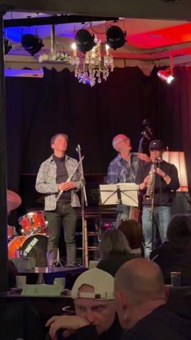 A jazz band plays on a raised stage in front of a seated audience. The band consists of a drummer playing a red Yamaha drum kit, a flutist dressed in black pants and a patterned shirt, a bass player wearing glasses and a plaid shirt, and a saxophone player in jeans and a black sweater. The stage is lit with blue and red lighting fixtures and a chandelier. The audience is seated at tables, with drinks visible. The band members are looking at each other, presumably coordinating the music. A microphone stands in front of each band member except the drummer.