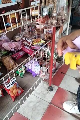 A person is in a store, holding snacks. The store has shelves stocked with various packaged food items, including chocolates, chips, and biscuits. The person is wearing dark pants and white sneakers. The floor is tiled with red and white squares. The video is captured from a high angle, looking down at the scene.