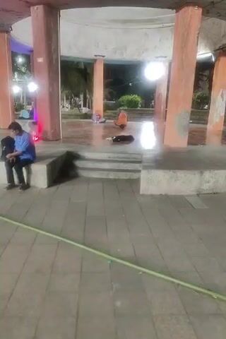 The video depicts an evening scene within a semi-circular, open-sided structure, likely a gazebo or bandstand, in a public park in Mumbai, India. The ground and the elevated platform within the structure are tiled and appear wet, suggesting recent rain or cleaning, with artificial lights reflecting on the shiny surface.

On the left side of the frame, a man in a blue shirt and dark pants is seated on a concrete ledge, looking down at a black backpack positioned next to him. On the main, elevated platform, a black and white dog is lying down, seemingly asleep or resting. As the video progresses, a man wearing an orange shirt and blue shorts walks across the wet platform. Another individual, dressed in a light-colored top and blue pants, is also seen sitting on the platform.

The structure is supported by thick, orange-brown painted pillars. Artificial lighting illuminates the area, indicating it is nighttime or late evening. A green pipe or hose is visible on the tiled ground in the foreground. The scene appears calm and relaxed. No visible text is present.