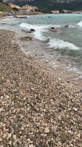 The video shows a rocky beach with waves gently rolling onto the shore. In the background, there are some buildings on a hillside and a body of water. The sky is overcast. The camera is stationary and focused on the beach and the water.