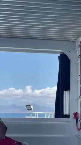 The video shows a view from inside a building or structure, looking out through a window. The window frame is white, and the ceiling above has a slatted design. Outside, there is a clear blue sky with fluffy white clouds. In the distance, there are mountains visible above a body of water, presumably the ocean or a large lake. On the balcony just outside the window, there's a spotlight mounted on a stand. Part of a dark blue curtain is visible to the right of the window. A person's head is briefly visible on the left side of the frame. There are faint sounds of people talking in the background.