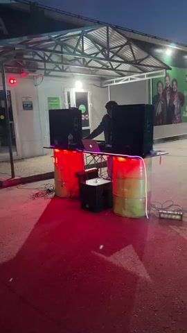 A person operates a DJ setup outdoors under a covered structure. The setup includes two large speakers, a laptop, and a smoke machine. The DJ booth is constructed with two barrels and a tabletop, illuminated by red and blue LED lights. The ground is paved and appears wet, reflecting the red lights. In the background, a building with windows and a mural featuring three women is visible. The sky is dark, indicating it is either dusk or night.