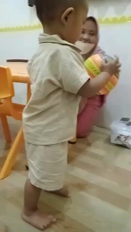 A toddler, wearing a light brown collared shirt and shorts, stands on a wooden floor and holds a colorful ball with orange, yellow, green, and blue sections. The toddler appears to be interacting with an adult woman kneeling behind a small orange table. The woman wears a pink hijab and pink clothing, and a light-colored mask covers her mouth and nose. Her hands are extended towards the toddler, and she is smiling. In the background, a white plastic container with a label displaying the number "7" is visible on the floor. The wall behind the woman has a yellow and white striped pattern. The room appears to be a child's play area or classroom.