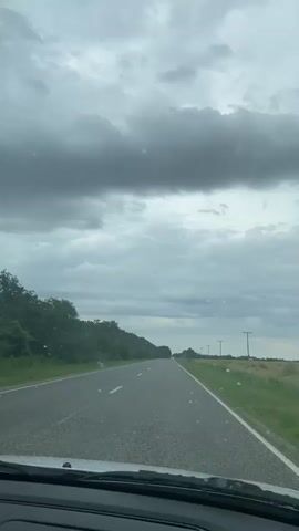 This is a first-person perspective video taken from inside a vehicle driving on a paved road. The road is empty of other vehicles and is lined with trees on the left and a field on the right. The sky is overcast with dark clouds. The timestamp indicates the video is only a few seconds long and shows continuous movement along the road.