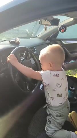 The video shows a toddler, viewed from the right side, standing in the driver's seat of a car. The toddler is wearing a white shirt with a pattern of cartoon animals and green pants with a car pattern. The toddler's hands are on the steering wheel, and their head is turned away from the camera. The car's interior is visible, including the dashboard, steering wheel, and part of the passenger seat. The car appears to be parked outdoors, with sunlight entering the vehicle, casting shadows. A small toy penguin is hanging from the rearview mirror.