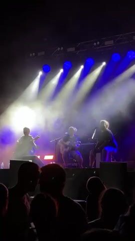 A live music performance is captured from a low angle, with the stage lit by bright blue and white spotlights. Three musicians are visible on stage. The musician on the left plays a stringed instrument that resembles a violin. The musician in the center plays a guitar. The musician on the right is seated and appears to be singing into a microphone. Smoke or haze is present on the stage, illuminated by the lights, creating beams of light. In the foreground, the silhouettes of an audience are visible. The overall atmosphere is that of an outdoor or large indoor concert.