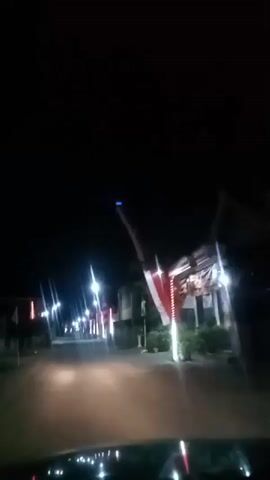 The video shows a dark outdoor scene at night, likely from a moving vehicle. The perspective is looking forward down a street illuminated by streetlights and decorative lights. On either side of the street, there are buildings and some greenery. Several Indonesian flags are visible, mounted on poles, adding a patriotic element to the scene. The road ahead is visible, and the vehicle's dashboard or hood is partially in the foreground, suggesting the footage is captured from inside a car. The overall atmosphere is calm and serene, with the lights creating a festive or celebratory feel.