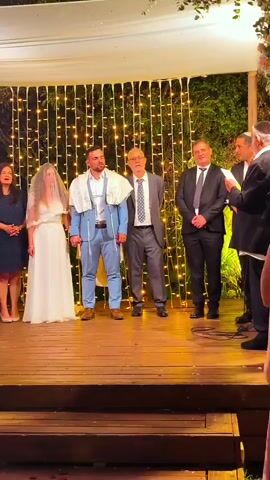 The video captures a wedding ceremony taking place outdoors under a canopy adorned with string lights. A bride and groom stand at the center of a wooden stage, facing a man in a suit and tie who appears to be officiating. The bride wears a white gown and a veil, while the groom is dressed in a light blue suit, also wearing a tallit. To their left, a woman in a dark blue dress stands. To the groom's right stand two men in suits. The officiant holds a booklet and seems to be speaking. The overall atmosphere appears formal and celebratory, with the string lights creating a festive backdrop. The video has a slight upward tilt as it progresses.