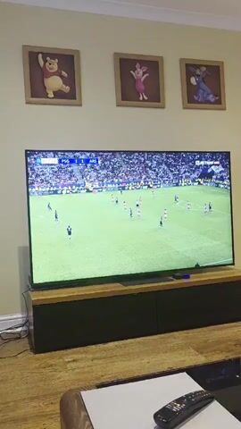 A wide shot of a large flat screen TV displaying a soccer game. The score is PSG 1 ARS 1. The television is resting on a dark entertainment center with a light wooden trim. Above the TV are three framed Winnie the Pooh characters: Pooh, Piglet, and Eeyore. The room has light tan walls and a light wood floor. A white desk with a TV remote rests in the lower right corner of the frame.