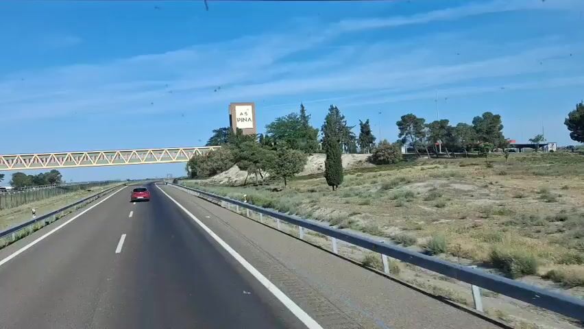 The video shows a view from inside a vehicle driving on a highway. The sky is clear and blue. On the right side of the road is a sign that says "A.S. PINA" on a tall, rectangular structure. A pedestrian bridge, painted green and brown, crosses over the highway. The road has multiple lanes and a guardrail. There are other cars on the road, including a red car traveling ahead. The surrounding landscape includes trees, shrubs, and a grassy area. The vehicle is moving forward, passing under the pedestrian bridge.