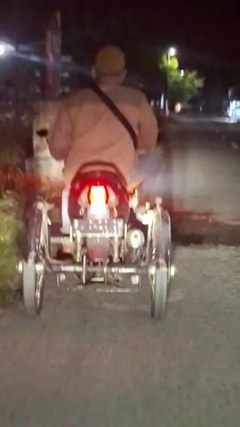 The video shows a man riding a small three-wheeled vehicle at night. The vehicle has a single rear light and a chrome frame. The man is wearing a light brown shirt, jeans, and a hat, with a messenger bag slung across his shoulder. He is driving down a paved road with streetlights and buildings visible in the background. The view is from behind the vehicle, showing its movement away from the camera.