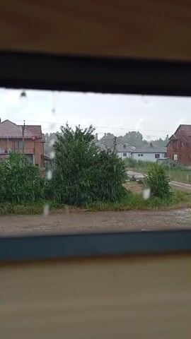 The video is a close-up shot looking through a window frame at an outdoor scene during a rainstorm. Raindrops are visible on the windowpane and streaking down, distorting the view of the environment outside. The scene outside includes houses with red roofs, lush green trees, and a dirt road. The sky appears overcast and cloudy, indicative of the rainy weather. There are no people visible in the frame.