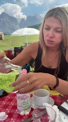 A woman with blonde hair and a black tank top is pouring water from a clear bottle into another bottle on a red and white checkered tablecloth. She is wearing a gold ring on her left ring finger and a pendant necklace. In the background, there are mountains, a white umbrella, chairs, and green grass. A white coffee cup and saucer are on the table.