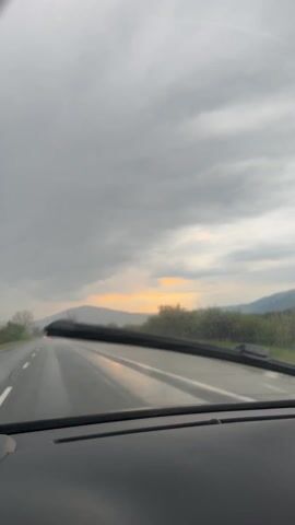 The video, filmed from inside a vehicle, captures a drive along a wet road during overcast weather, likely in Konare, Bulgaria. The windshield wipers are actively clearing rain from the windscreen. The sky is filled with dark, heavy clouds, but a strip of yellowish light is visible on the horizon, suggesting either sunrise or sunset. Mountains are visible in the distance, partially obscured by the low-hanging clouds. A white vehicle, possibly a van or truck, is driving ahead in the same direction. The sides of the road feature green fields and scattered trees and bushes, some appearing bare. The overall scene depicts a journey on a rainy, grey day with a hint of warm light breaking through the clouds. No people or visible text are present.
