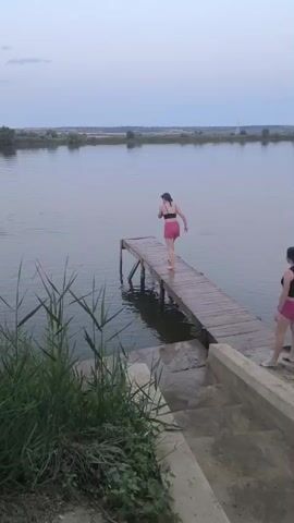This media file shows a serene natural scene at dusk in Ciumai, Moldova. The main subject is a young woman in a black top and pink shorts, standing at the end of a wooden pier that extends into a calm body of water. The pier itself is weathered and shows signs of age, with visible support beams submerged in the water.

The woman is captured mid-jump as she leaps off the pier into the water. The splash as she enters the water creates a dynamic visual element against the otherwise still surface. Another person, also wearing a black top and pink shorts, is visible on the pier closer to the shore, watching the jumper.

The setting is a lakeside or riverfront with lush green reeds and grass in the foreground. The water is a muted, greyish-blue, reflecting the soft, overcast sky. The background features a low, tree-lined horizon with some distant structures, possibly indicating a rural or semi-rural area. The overall atmosphere is peaceful and natural, with the muted colors and soft light suggesting late evening. There is no visible text in the media file. The activity is recreational, with the individuals enjoying a swim on what appears to be a calm, possibly warm evening.