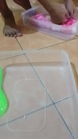 A person's bare feet are visible on a tiled floor. The person is crouching over a clear plastic container holding a pink and white object, possibly fabric or food. Their hands are submerged in the container, manipulating the object. In the foreground, another clear plastic container is visible with a green, bulbous object inside.