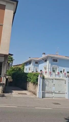 This video shows a blue building with a white trim and a terracotta roof. There are large double gates in front of the building with the letters B&B on them. The building has several windows and balconies, some of which are decorated with pink flowers and small topiaries. There are trees and bushes surrounding the property. The sky is clear and blue. The video is shot from a moving vehicle, likely a car or motorcycle, as indicated by the slight camera shake and the perspective.