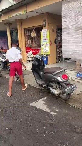 The video shows a young boy wearing a white shirt and red shorts, walking away from the camera towards a small shop. He has a black cap on and is wearing flip-flops. He is walking past a pink bicycle and a black motorcycle. The shop has a sign in yellow with text that appears to be in Indonesian, and it lists various payment methods. There are also ice cream freezers visible inside the shop. The ground is wet in some areas, suggesting it may have recently rained. The overall environment appears to be a street in a residential or commercial area.
