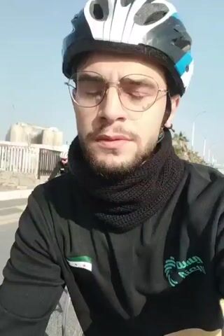 The video shows a group of people riding bicycles on a sunny day. The person filming the video is among the cyclists, wearing a helmet, glasses, and a black scarf. They are dressed in a black long-sleeved shirt with a small green and white flag on the sleeve. Other cyclists are visible in the background, many also wearing helmets. Palm trees line the side of the road, and a few cars are visible in the distance. The group appears to be cycling together along a paved road.