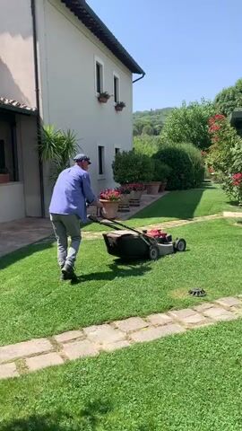A man pushes a red and black lawnmower across a green lawn. He is wearing a blue shirt, grey pants, and a blue cap. He is walking from left to right across the frame. Behind him, there is a light-colored house with windows and potted plants. To his right, there is a green gazebo with tables and chairs. The lawn is bordered by a stone pathway and surrounded by trees and bushes. The sky is blue and clear.