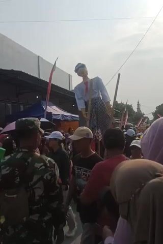 This is a video taken from a ground-level perspective at an outdoor event. The focal point is a very large effigy of a person dressed in a white shirt, red pants, a white and black checkered sarong, and a blue and white head covering. The effigy is mounted on two tall wooden poles and appears to be held upright by several people, including individuals wearing camouflage uniforms, a person in a bright green safety vest, and others in civilian clothing. The effigy's head is tilted back. A crowd of people, many wearing headscarves, is gathered around, watching the effigy. Tents and temporary structures are visible in the background, along with flags and the silhouette of a building with a minaret. The lighting suggests it is daytime.