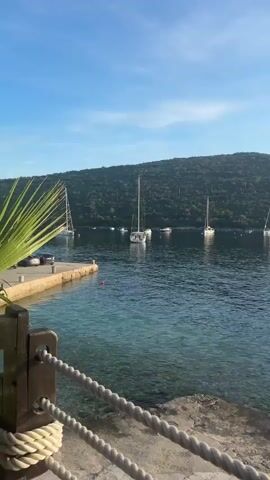 The video captures a serene coastal scene in Bigova, Montenegro, transitioning from a daytime view to a golden hour glow. Several sailboats and catamarans are anchored in the calm, clear blue waters of a bay. The bay is framed by lush, green, tree-covered hills under a partly cloudy, blue sky.

In the initial shot, the camera is positioned low, looking out over a stone jetty with a rope barrier in the foreground, towards the anchored boats and the distant hills. As the video progresses, the camera pans to the right, revealing the sun beginning to set, casting a bright, shimmering reflection across the water. Two rustic straw sun umbrellas are visible on a small beach or pier in the foreground, with a person partially visible near one. The scene evokes a peaceful, relaxing atmosphere, typical of a coastal resort or quiet cove. There is no visible text.