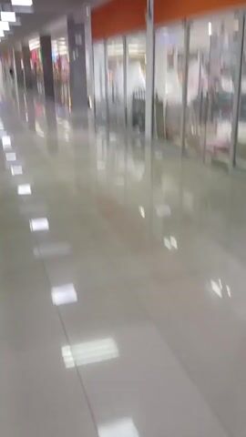 The video shows the interior of a commercial space, likely a shopping mall, from a low angle. The floor is tiled and reflective, showing the reflection of the overhead lighting fixtures. The ceiling is a grid pattern with recessed lighting. The corridor is lined with storefronts, some with glass displays. The stores appear closed or empty. The overall impression is of a clean, modern, and somewhat deserted space. No people are visible.