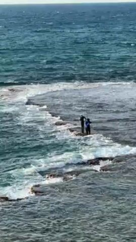 Two people stand on a rocky outcrop in the ocean. Waves crash around them. The water is a deep teal color. The sky is a pale blue.