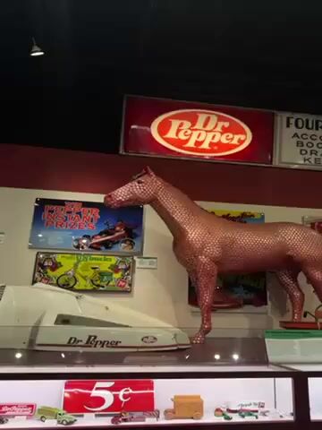 The camera pans up to focus on a life-size horse sculpture covered in what appear to be Dr. Pepper bottle caps. The sculpture is reddish-brown in color and positioned within a Dr. Pepper museum or display. Behind it is a Dr. Pepper sign, while other Dr. Pepper-related promotional items and displays are visible in the surrounding environment.