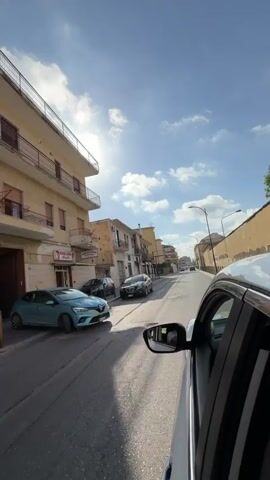 The video provides a first-person perspective from inside a white car driving along a street in Maddaloni, Italy, on a bright, sunny day with scattered white clouds. The camera is positioned to view the road ahead, capturing the car's right side and side mirror.

The street is lined with multi-story buildings, some appearing residential and others housing ground-floor businesses. Visible signs on the left side of the street include "MACELLERIA" (Butcher shop) and "BISTECCHERIA" below it, and further down, "PIZZERIA." The buildings, some in light yellow and others beige or white, feature balconies and show signs of wear.

Several cars are visible on the street: a black car and a light blue car are parked on the left side of the road, while a small blue car and another white car are seen driving in the opposite direction further down the street. The sun is high in the sky, casting strong light and shadows. A person's arm and hand are visible in the reflection of the car's side mirror. The asphalt road surface appears worn.