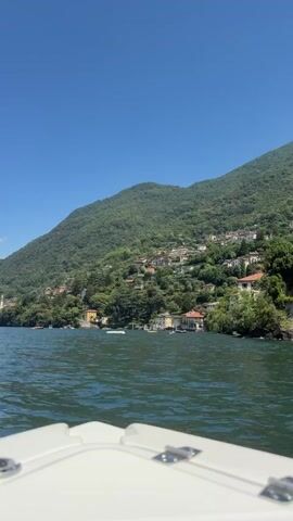 The video shows a view from a boat on a lake. In the foreground, the white bow of the boat is visible. The lake water is a deep green and reflects the clear blue sky. In the background, a lush green, tree-covered mountain rises steeply from the lake. Houses with red tile roofs are nestled along the mountainside, appearing to cling to the slopes. Several small boats are visible on the water, including a white jet ski leaving a spray in its wake. The overall scene is one of a sunny day in a scenic, possibly European, lakeside setting.
