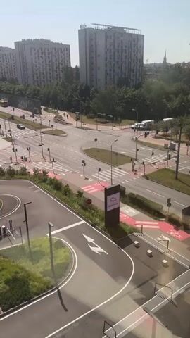 The video shows an aerial view of a busy intersection on a sunny day. Tall apartment buildings and trees are visible in the background. The foreground shows a circular road with arrows indicating the direction of travel, and a pedestrian walkway. A tall, rectangular sign with a pink logo is located near the walkway. Cars are seen driving through the intersection. Traffic lights and pedestrian crossings are present. The overall scene depicts an urban environment with moderate traffic.