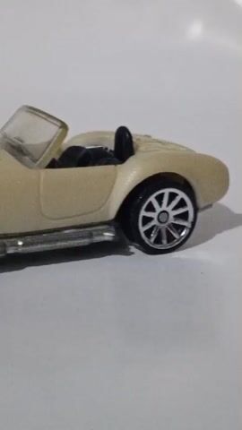 The video shows a close-up, side-view, panning shot of a tan toy car. The car is a convertible with the top down and has chrome-colored side exhaust pipes. The wheels are silver with a multi-spoke design. There are some yellow, flame-like graphics on the hood of the car. The car is on a plain white surface.