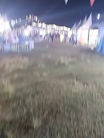 The video shows a nighttime scene at what appears to be a fair or carnival. The camera pans across a grassy field with several carnival rides illuminated by bright lights. String lights are strung above, and colorful pennant banners flutter in the night sky. Several food or game booths are set up under tents, and people can be seen indistinctly. The overall atmosphere is festive and lively.