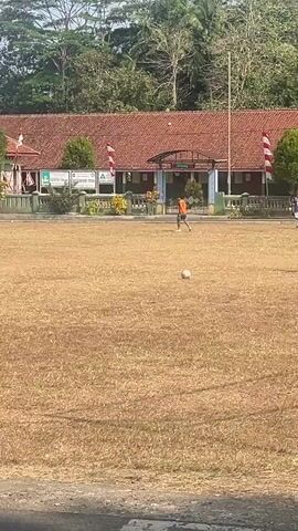 A soccer ball is in motion on a dry, brown grass field. A person in an orange shirt and black shorts is kicking the ball. Several other people in similar attire are scattered across the field. In the background, a building with a red tile roof and white trim stands under a bright, clear sky. Trees, including palm trees, are visible behind the building. The scene appears to be a sports practice or game taking place outdoors.