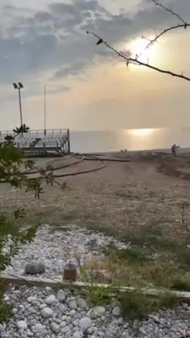 The video shows a beach scene at sunset. A viewing platform is visible on the left side of the frame. The sun is shining brightly, reflecting on the sea. Some people are walking on the beach. The foreground includes a stony area with some greenery. The camera moves slightly, focusing more on a bush.