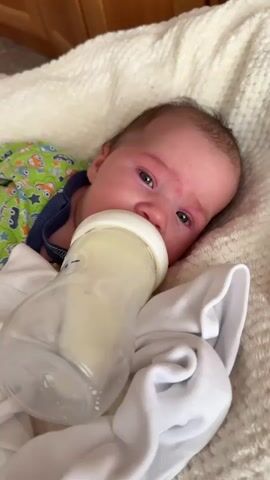 A baby lies on its back on a cream-colored textured blanket. The baby is wearing a green onesie with a repeating pattern of blue and orange cartoon aliens and green stars. The baby's head is turned slightly to its left, and its eyes are open, looking towards the camera. A clear plastic baby bottle with a white nipple is positioned to the baby's mouth, and the baby appears to be drinking from it. The bottle contains a light yellow liquid. A white cloth is draped over the bottle and the blanket. The background shows a wooden surface.