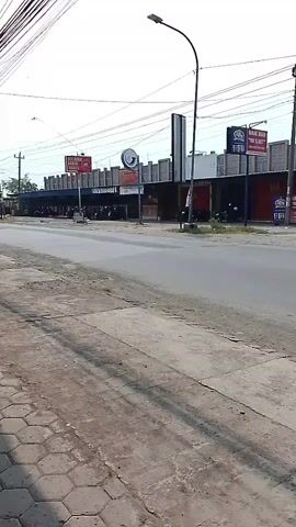 The video shows a street scene with several vehicles moving. In the foreground, there is a paved sidewalk with a hexagonal pattern. The road is visible, with a dark gray surface. On the other side of the road, there is a row of buildings with signs. One sign is red and has text in what appears to be an Asian language. Another sign is round with a circular logo. There are also multiple signs with "FBI" written on them. Several motorcycles are parked in front of the buildings.

In terms of actions, a person riding a motorcycle with a passenger is seen traveling from left to right across the frame. A black pickup truck is also seen traveling in the same direction. Further down the road, a red truck is observed moving from left to right. The sky is overcast, suggesting a daytime scene. The environment appears to be an urban or semi-urban area with overhead wires strung across the scene.