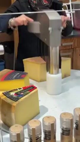 A person is shown using a stainless steel cheese shaving tool to cut a piece of cheese for a sample. The cheese shaver has a long cylindrical base and a lever arm. On the table in front of the machine are several wedges of cheese labeled with different names and a row of small jars labeled "Planfin". The background is blurry.
