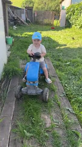 A toddler wearing a blue cap, a light-colored t-shirt, and gray shorts sits on a blue toy tractor on a wooden pathway surrounded by grass. The toddler has their hands near their mouth and appears to be looking at the camera. The background shows a wooden fence, a building, and a grassy hill under a clear sky. The video is shot from a low angle, focusing on the child and the toy.