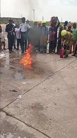 Here is a concise description of the scene:

The scene appears to be a fire safety demonstration taking place outdoors on a concrete surface. A small fire is burning on the ground in front of a group of people, including firefighters, police officers, and civilians. A firefighter in full gear is using a fire extinguisher to put out the flames. The audience watches intently, with some individuals recording the demonstration. The environment is open with buildings visible in the distance, under a slightly overcast sky. The atmosphere suggests an educational and practical session on how to handle small fires.