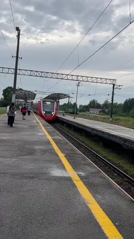 A modern, red and white train approaches a platform at a train station. The train has its headlights on and is labeled with the number 3C104-034. Several people are standing on the platform, waiting for the train. The platform is paved with asphalt and has a yellow line marking the safety zone. Overhead power lines and supporting structures are visible. The sky is overcast. The train appears to be moving at a moderate speed.