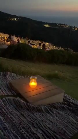 A low-angle video shows a wooden table with a lit candle on it, placed on a woven mat on a grassy slope at dusk. In the background, a group of people are seated at tables on a higher elevation, with lights from a town dotting a hillside overlooking the ocean. The sky displays a gradient of colors from deep blue to orange and pink at the horizon. The camera pans slightly to the right, revealing a person from behind, wearing a patterned shirt, looking out at the scenic view. The overall atmosphere is serene and romantic.