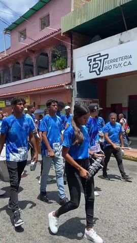 A group of young people walk along a street, playing music. They are dressed in matching blue t-shirts with a unique design. Some are carrying and drumming on large snare drums, while others have smaller percussion instruments. They walk together, forming a procession. Behind them is a hardware store called "FERRETERIA RIO TUY, C.A." The buildings around them are colorful, and the weather seems sunny.